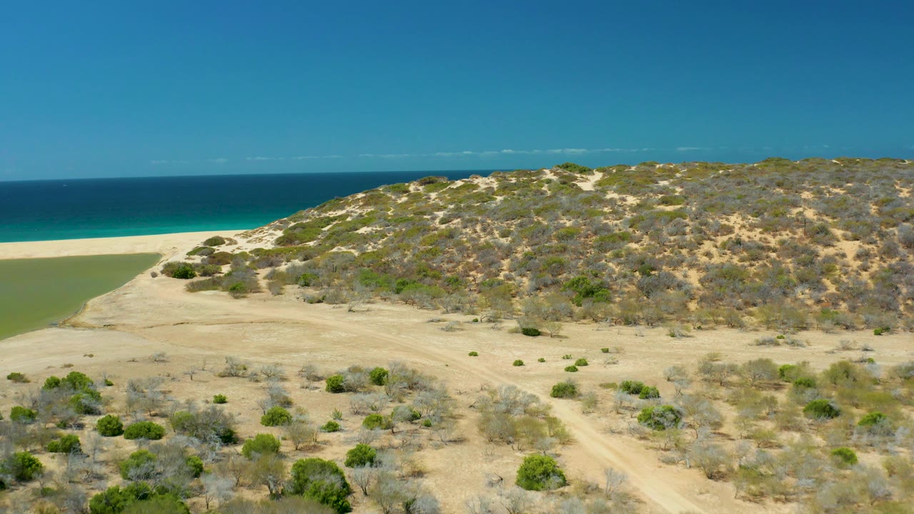 Wide drone view of arid dunes, beach, green lagoon water, and dark blue ocean.