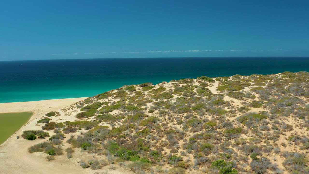 Aerial view over La Laguna Beach Los Cabos with scrub-covered sand dunes in front, a bright turquoise shoreline behind them, darker water near the horizon, and no visible people.