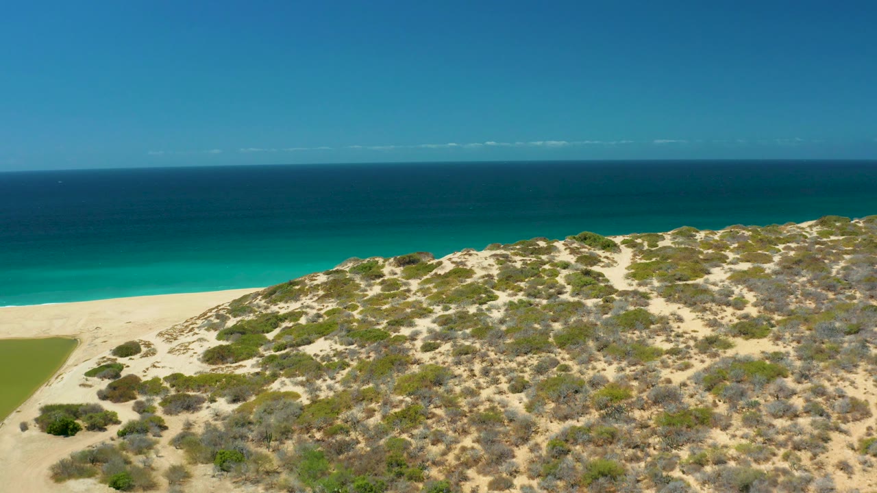 La Laguna Beach Los Cabos is seen in a wide aerial composition with sandy dune scrub across the lower frame, a pale beach strip, calm turquoise water, deep blue ocean, and no visible people.