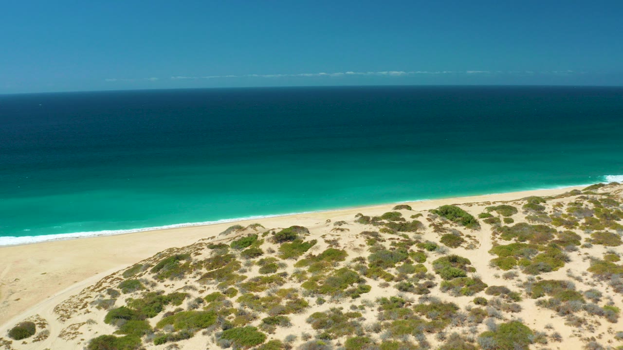 Aerial view over La Laguna Beach Los Cabos with shrub-covered dunes in the lower frame, pale sand at the shore, turquoise ocean beyond, and no visible people.