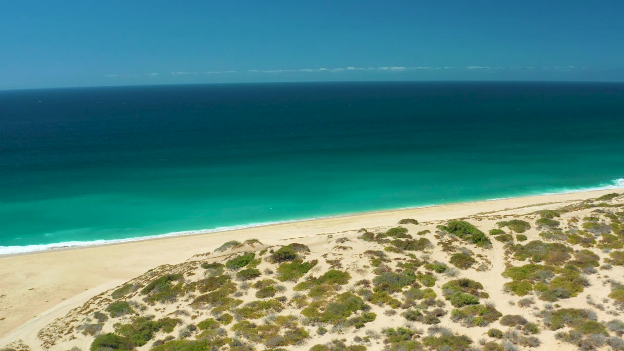 A high aerial frame of La Laguna Beach Los Cabos shows patchy dune vegetation, a clean sand strip, bright turquoise shallows, darker offshore water, and no visible people.