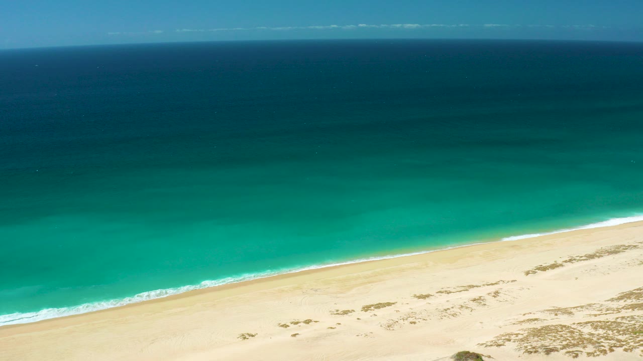 Aerial frame over La Laguna Beach in Los Cabos, Mexico, with deep blue water at the top, turquoise shallows near shore, pale sand below, sparse vegetation, and no visible people.