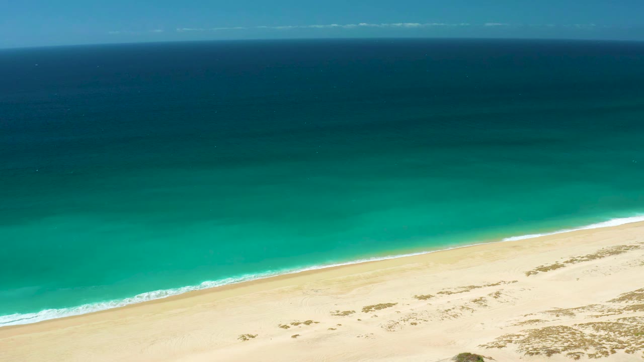 Aerial view at La Laguna Beach with the shoreline angled across the frame, turquoise water pressing toward pale sand, sparse beach vegetation, and no visible people.