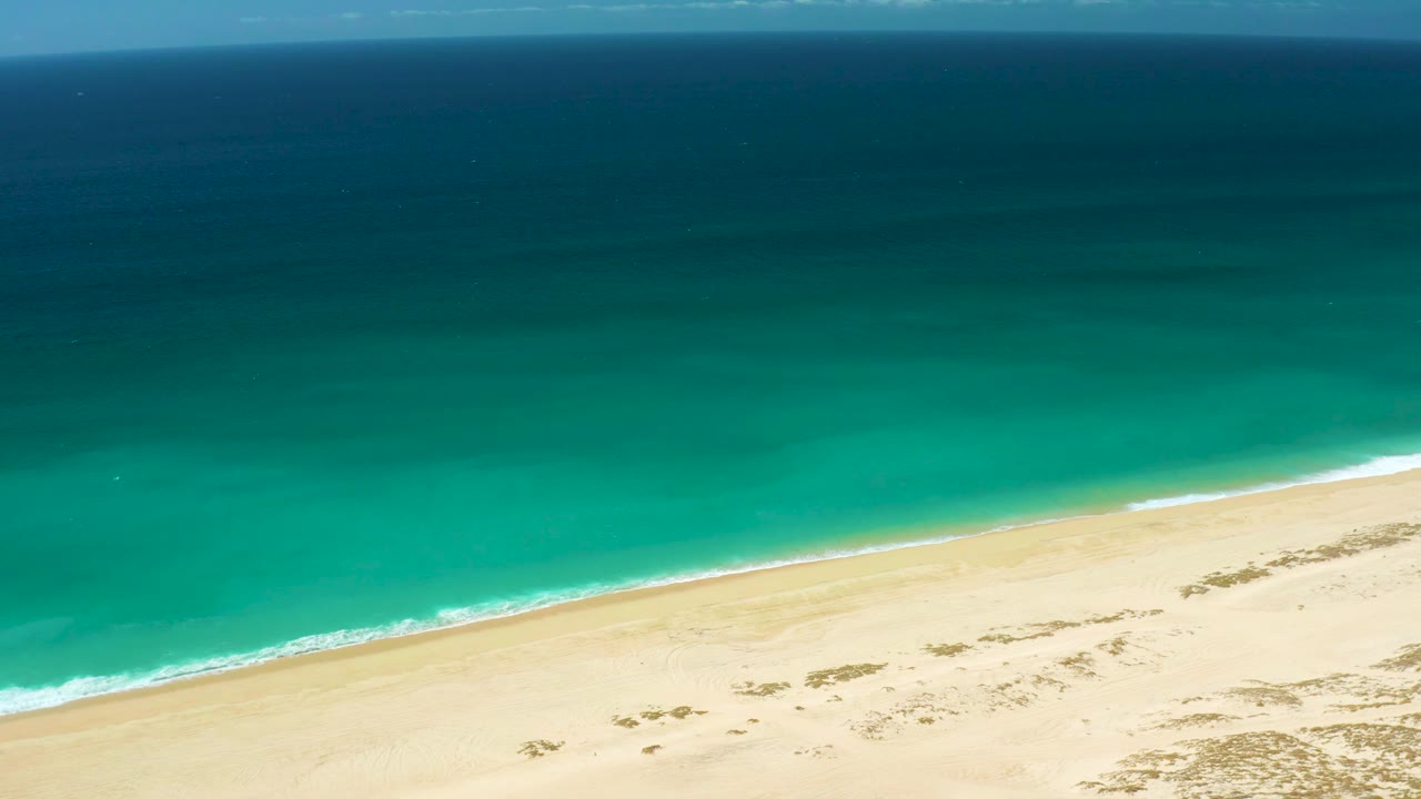 High aerial frame over La Laguna Beach showing darker offshore water, a broad turquoise band, a thin white break at the beach, pale sand, and no visible people.
