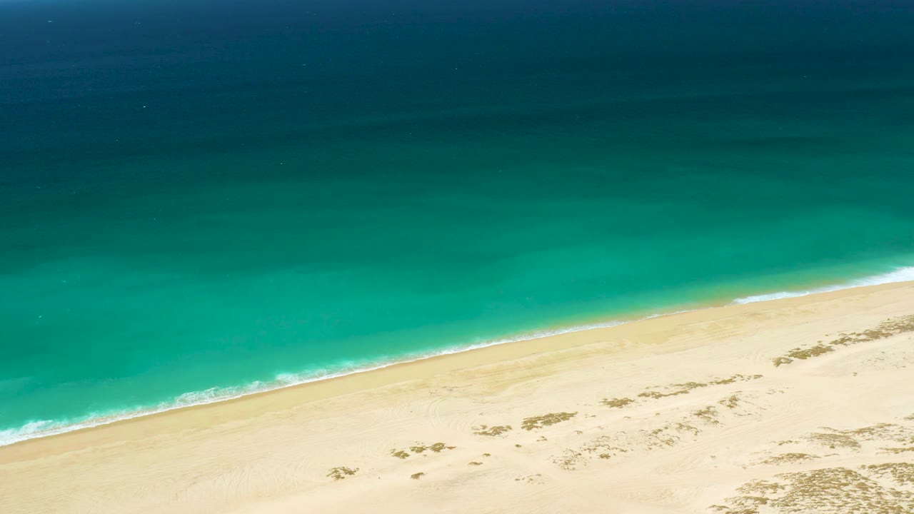 Aerial view of La Laguna Beach with ocean occupying most of the frame, a narrow white wave line along the diagonal shore, pale sand at the bottom, and no visible people.