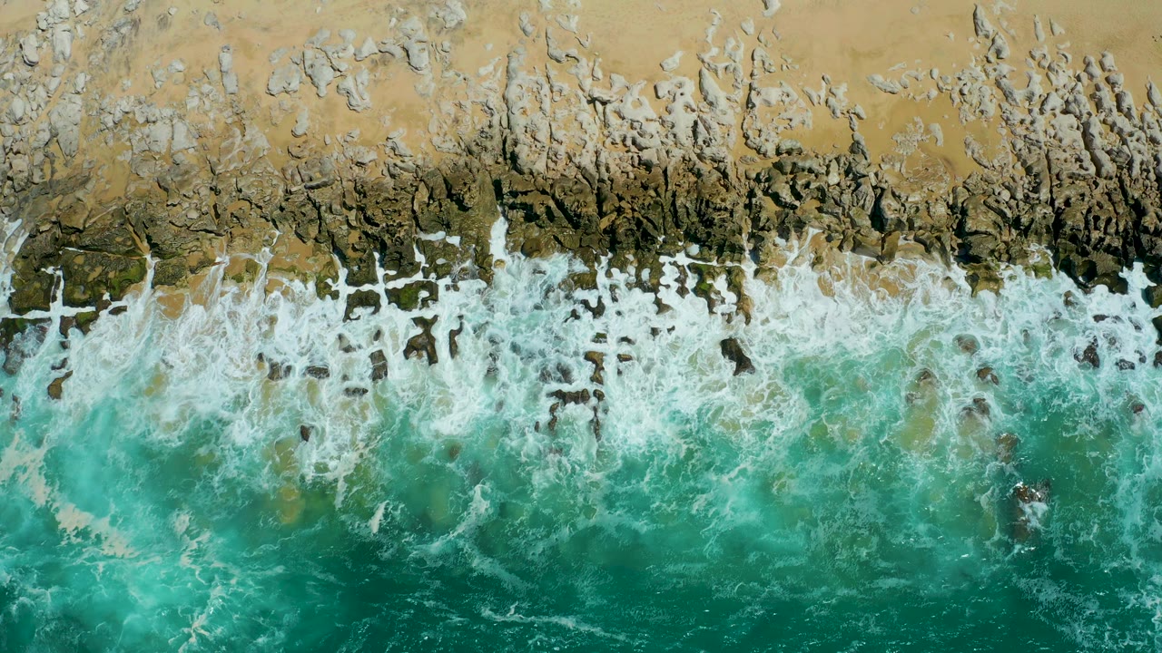 A top-down La Laguna Beach frame shows more broken whitewater over dark nearshore rocks, with turquoise water below and no visible people.