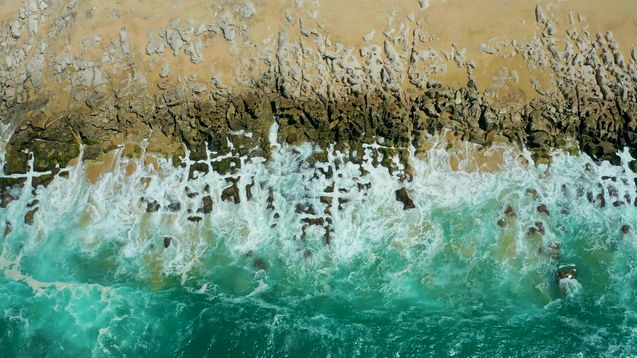 Overhead aerial view of a foamy wave line stretching across La Laguna Beach, with pale sand above, teal water below, and no visible people.
