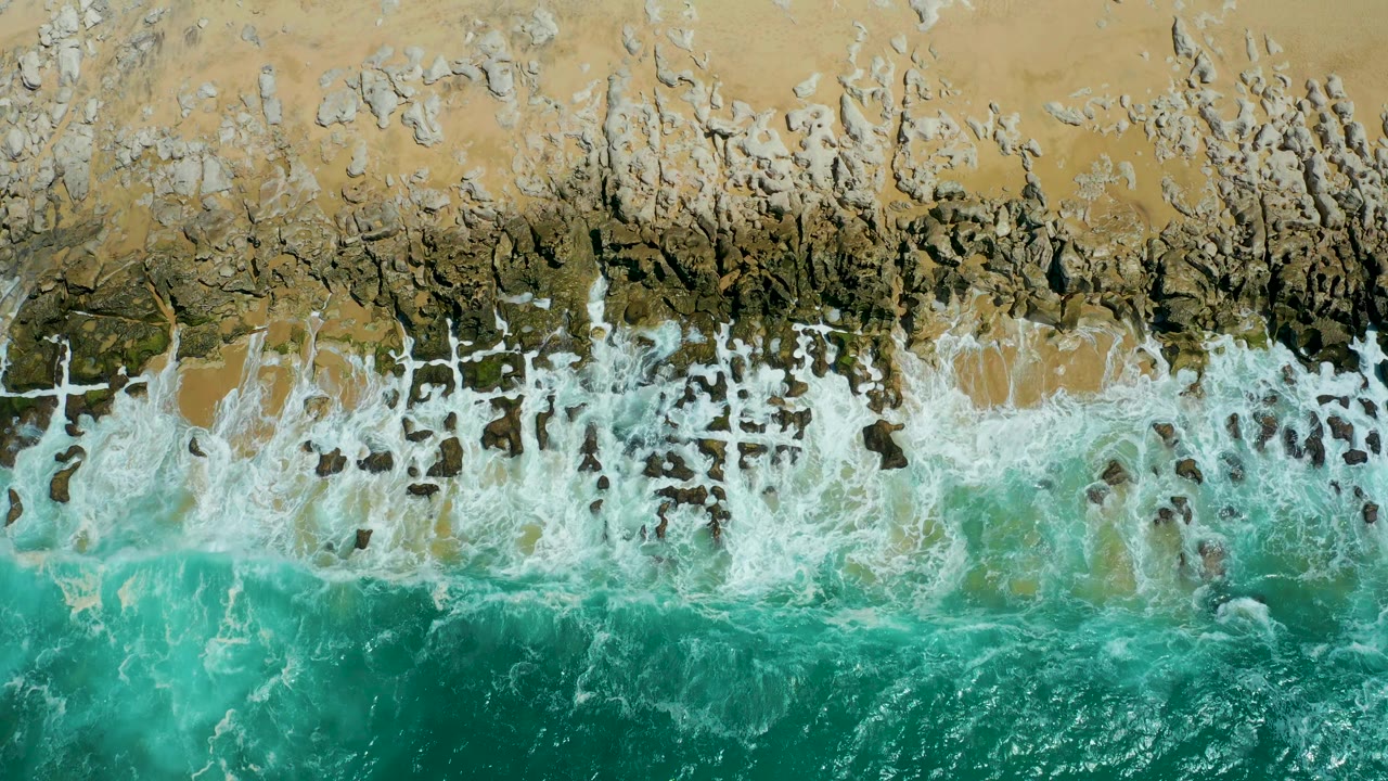 Top-down La Laguna Beach surf shows white foam gathering along the sand and rock edge while clear turquoise water fills the lower frame, with no visible people.