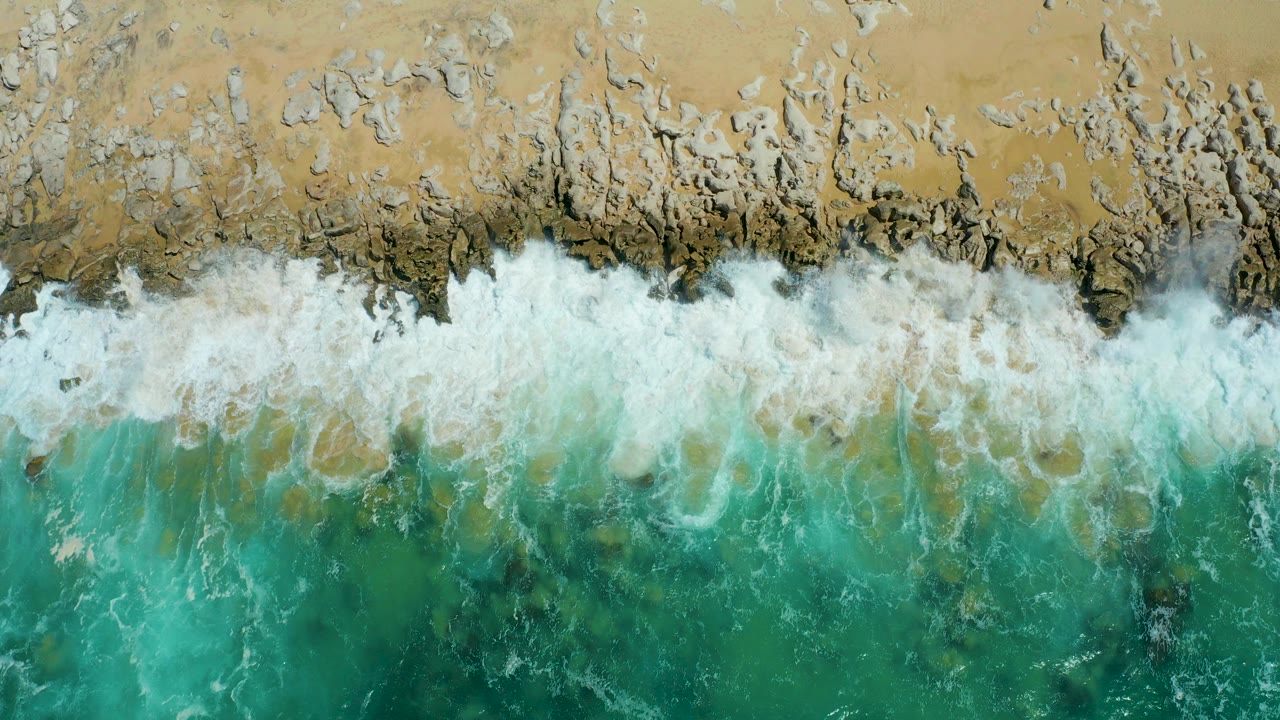 Top-down view of La Laguna Beach Los Cabos with turquoise water meeting a white foam line over rocks and tan sand, with no visible people.