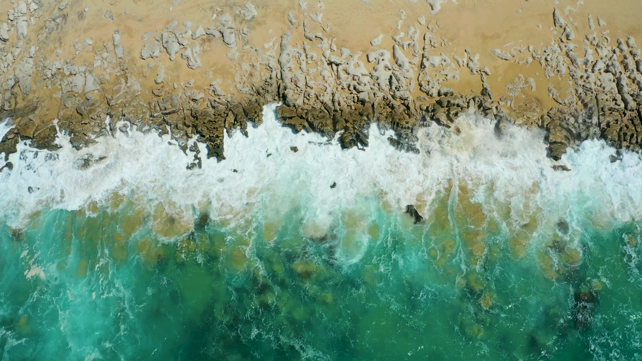 Top-down shoreline frame from La Laguna Beach Los Cabos showing turquoise water below a broken white surf line and pale sand above, with no visible people.