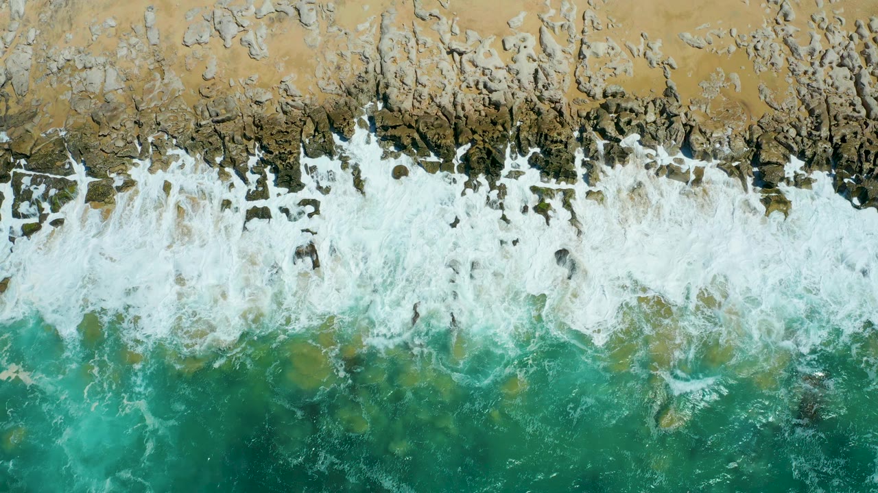 Aerial overhead frame at La Laguna Beach Los Cabos with white surf spreading wider across the rock band and turquoise water below, with no visible people.