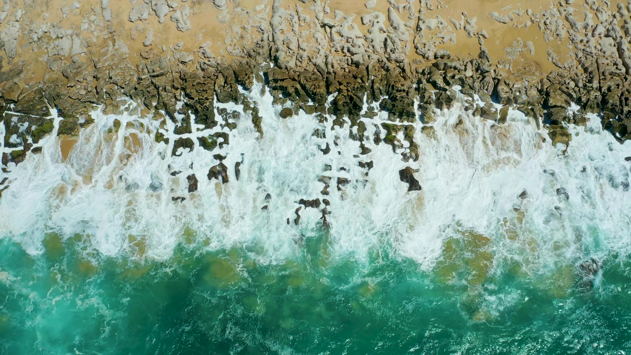 Overhead view of La Laguna Beach Los Cabos as thicker white surf covers much of the dark rocky strip below the tan beach, with no visible people.