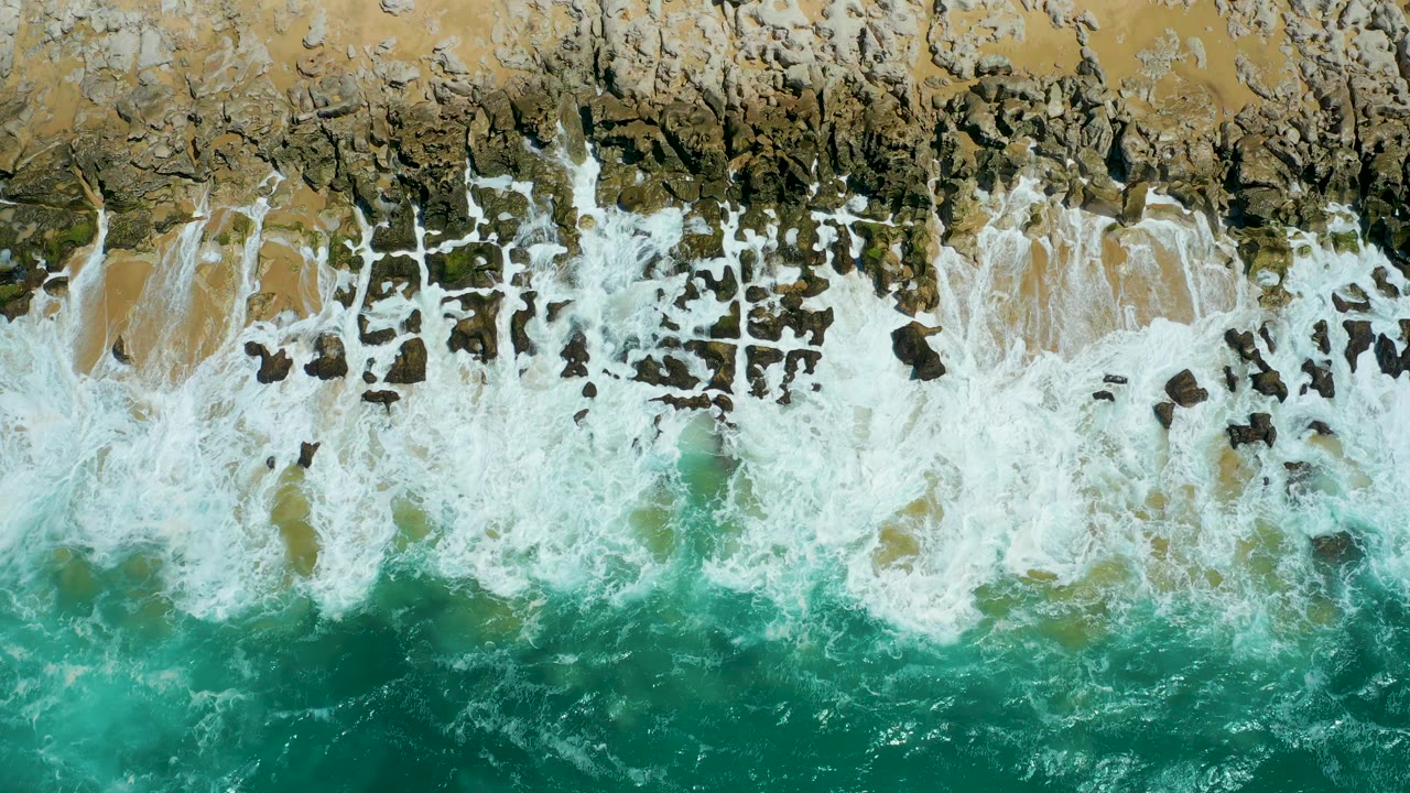 At 0:01, whitewater spreads farther across the dark rocky band at La Laguna Beach Los Cabos while turquoise water fills the lower frame, with no visible people.