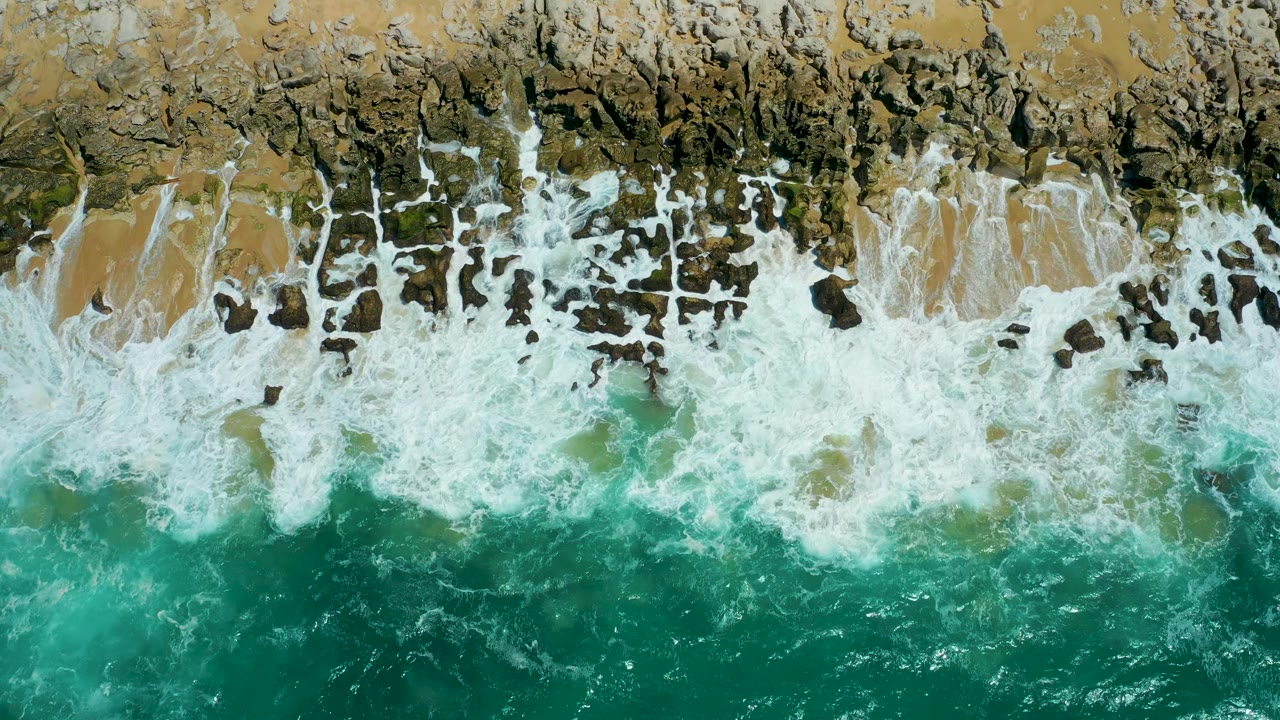 At 0:03, dense white foam covers much of the rocky and sandy edge at La Laguna Beach Los Cabos, with darker turquoise water below and no visible people.