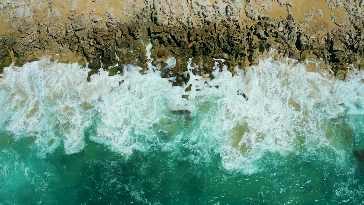 Top-down aerial view of white surf spreading across tan and dark rocks at La Laguna Beach Los Cabos, with turquoise water below and no visible people.
