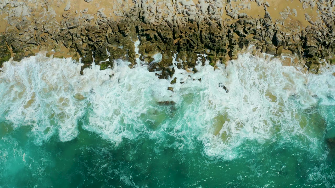 White ocean foam advances over dark rock patches and tan shore at La Laguna Beach Los Cabos, with clear turquoise water filling the lower frame and no visible people.