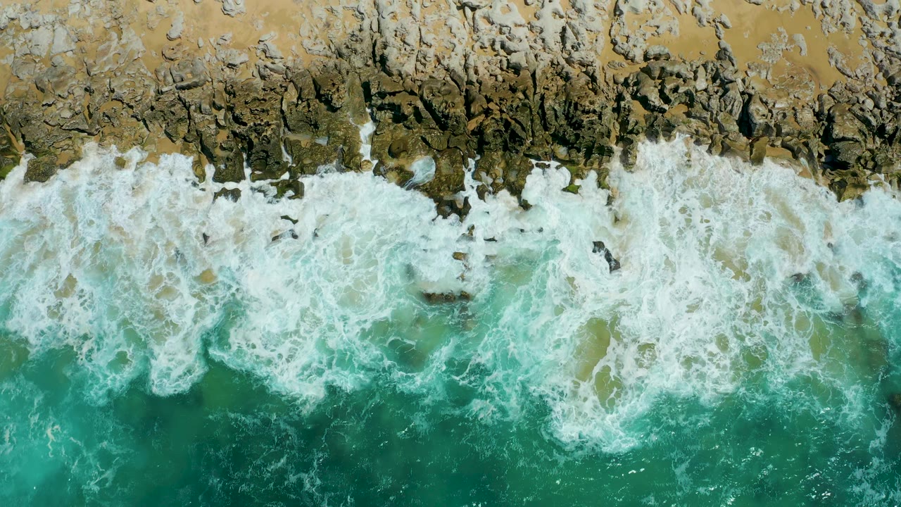A foamy wave band curls against a jagged tan and brown rocky edge at La Laguna Beach Los Cabos, with no horizon, structures, or visible people.