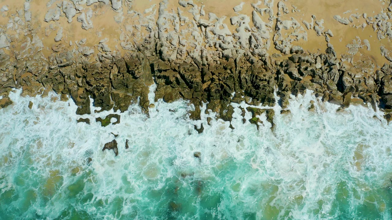 At La Laguna Beach Los Cabos, white surf spreads across dark shoreline rocks below tan sand, with turquoise water at the lower edge and no visible people.