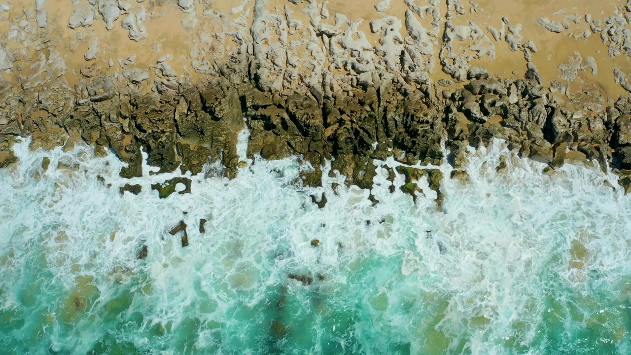 At La Laguna Beach Los Cabos, foamy turquoise surf churns below a narrow strip of tan sand and broken dark rock, with no visible people or structures.