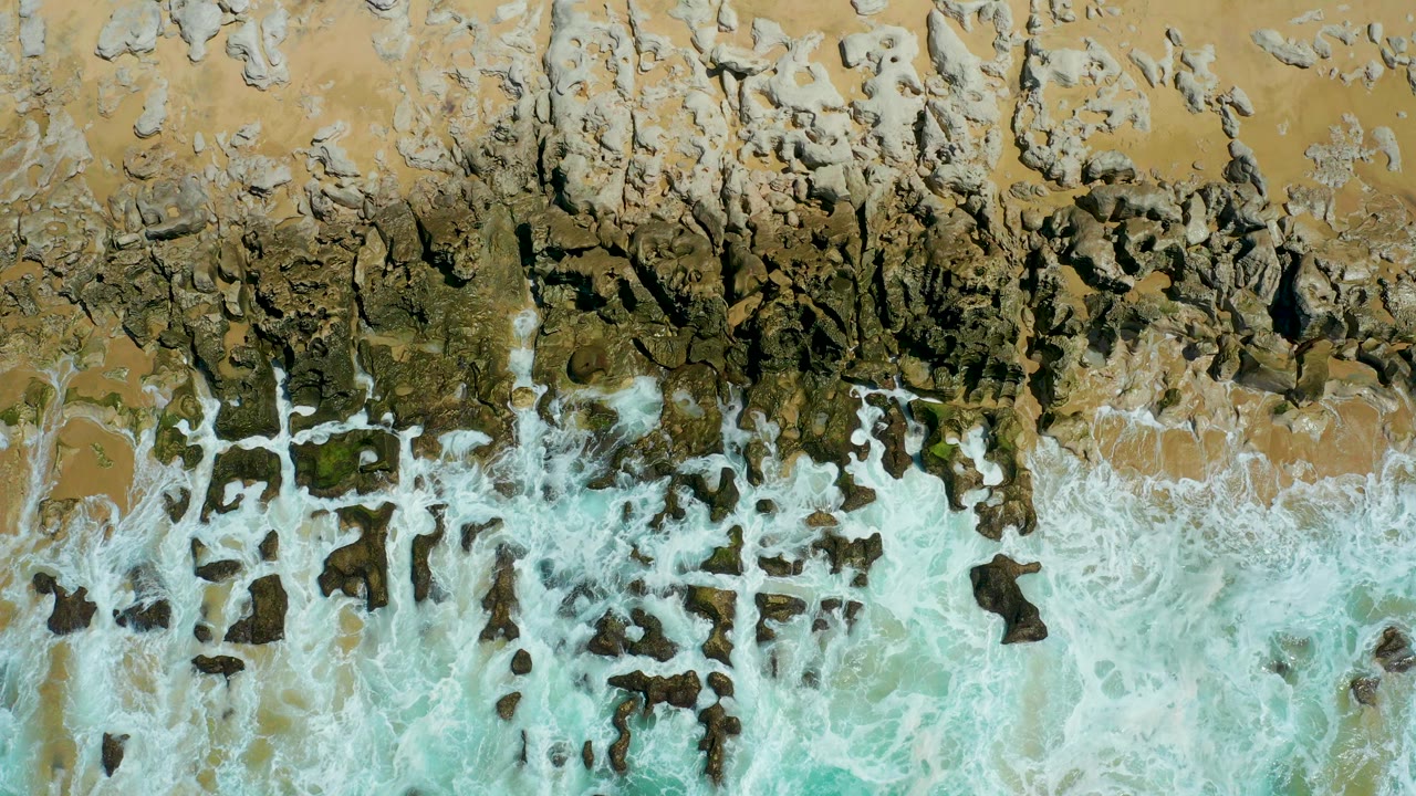 Overhead view of La Laguna Beach in Los Cabos with dark rock shelves, tan sand, turquoise water, and white foam entering the lower frame, no visible people.