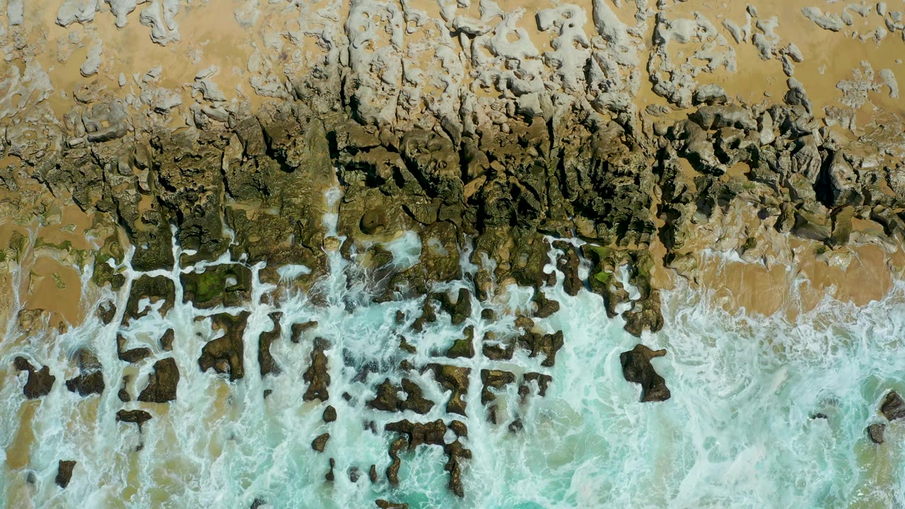 Top-down La Laguna Beach view with jagged rocks across the upper frame and pale turquoise surf breaking into white foam below, no visible people.