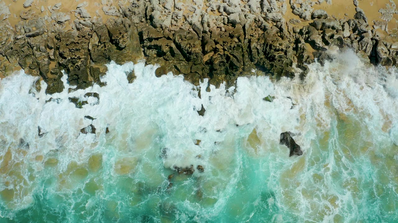 Overhead view at La Laguna Beach Los Cabos with a broad band of whitewater spreading across uneven rocks and shallow turquoise water, no visible people.