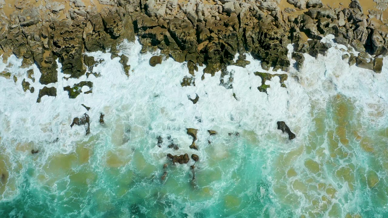 Top-down La Laguna Beach Los Cabos view with white foam curling around exposed dark rocks and green-blue water below the breaking line, no visible people.