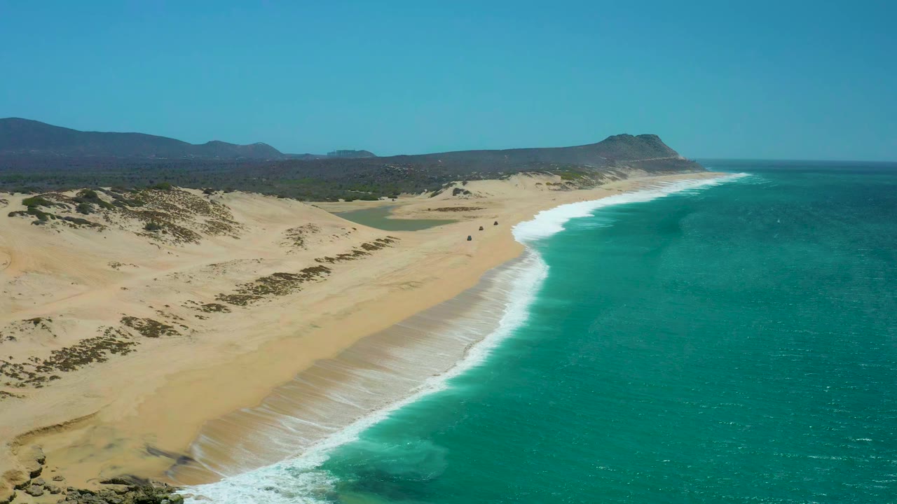 Aerial view at La Laguna Beach Los Cabos with the shoreline running diagonally, pale dunes inland, bright turquoise water offshore, distant mountains, and no visible people.