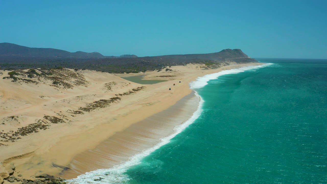 Aerial frame over La Laguna Beach Los Cabos showing more dune texture in the foreground, a long white breaker line along the beach, turquoise water, mountains on the horizon, and no visible people.