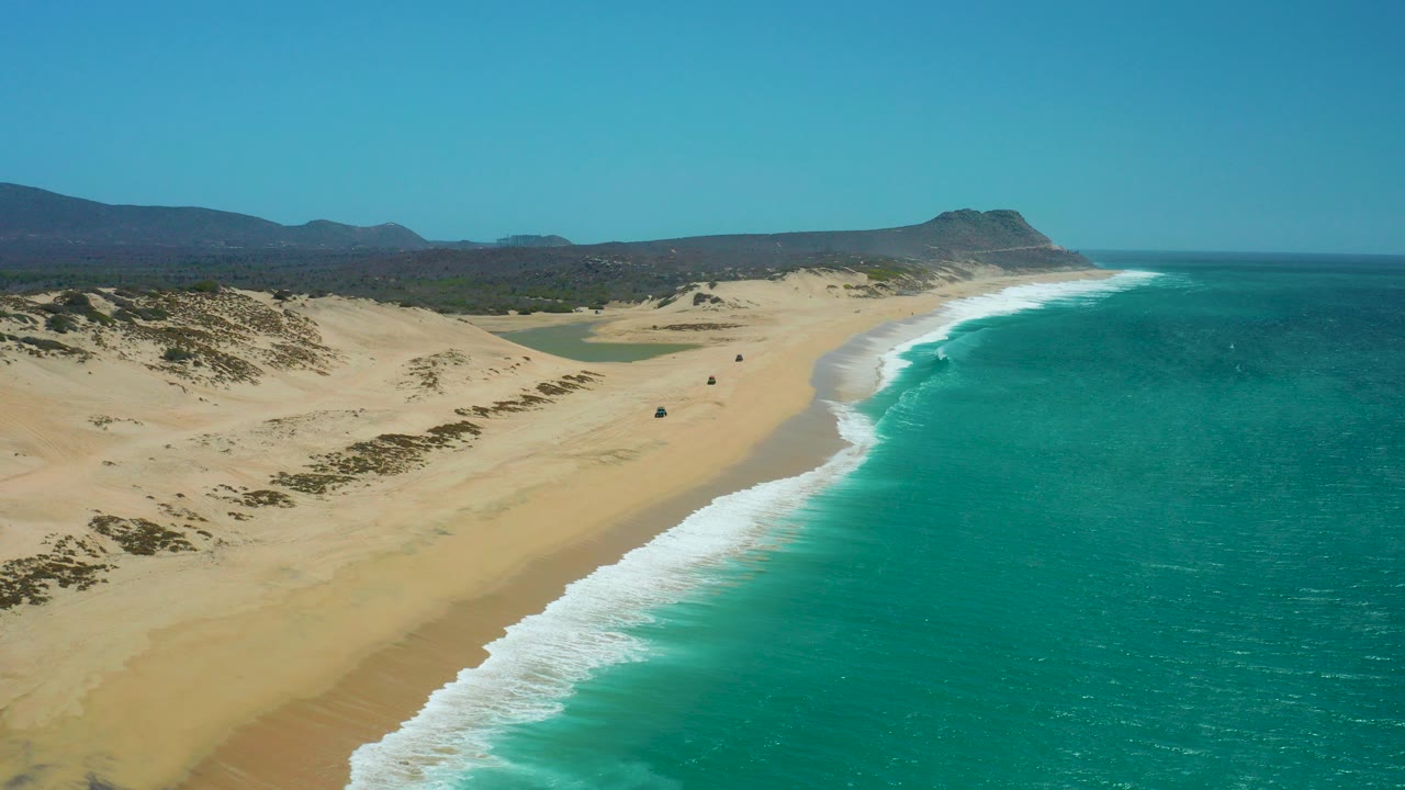 Opening aerial view of La Laguna Beach Los Cabos with turquoise ocean on the right, pale dunes on the left, distant hills beyond the shore, a small vehicle on the sand, and no visible people.