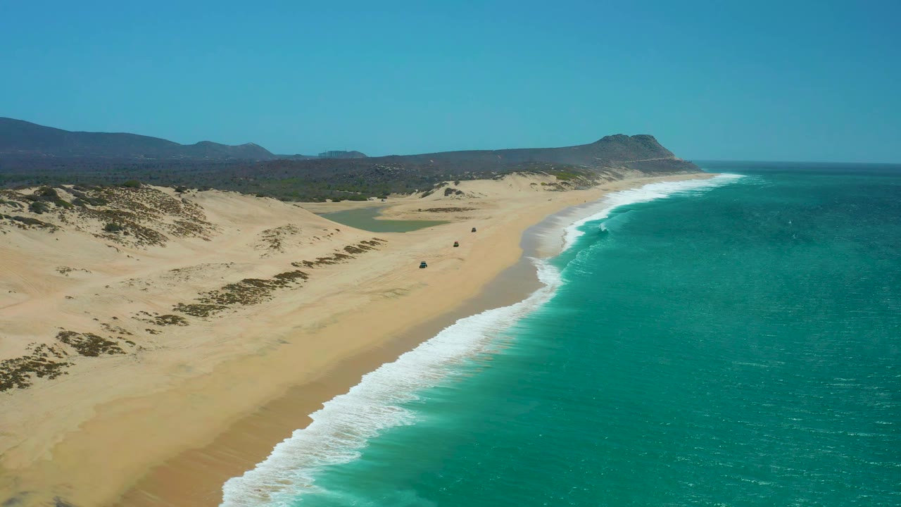 Wide aerial coastline with white waves, dunes, hills, and a small vehicle farther along the beach.