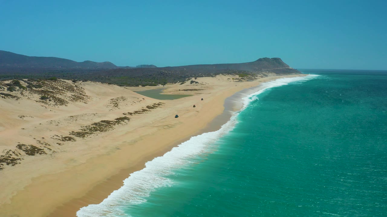 La Laguna Beach Los Cabos appears in a high coastal view with the shoreline curving toward distant hills, bright turquoise water, dune ridges, small vehicles on the sand, and no visible people.