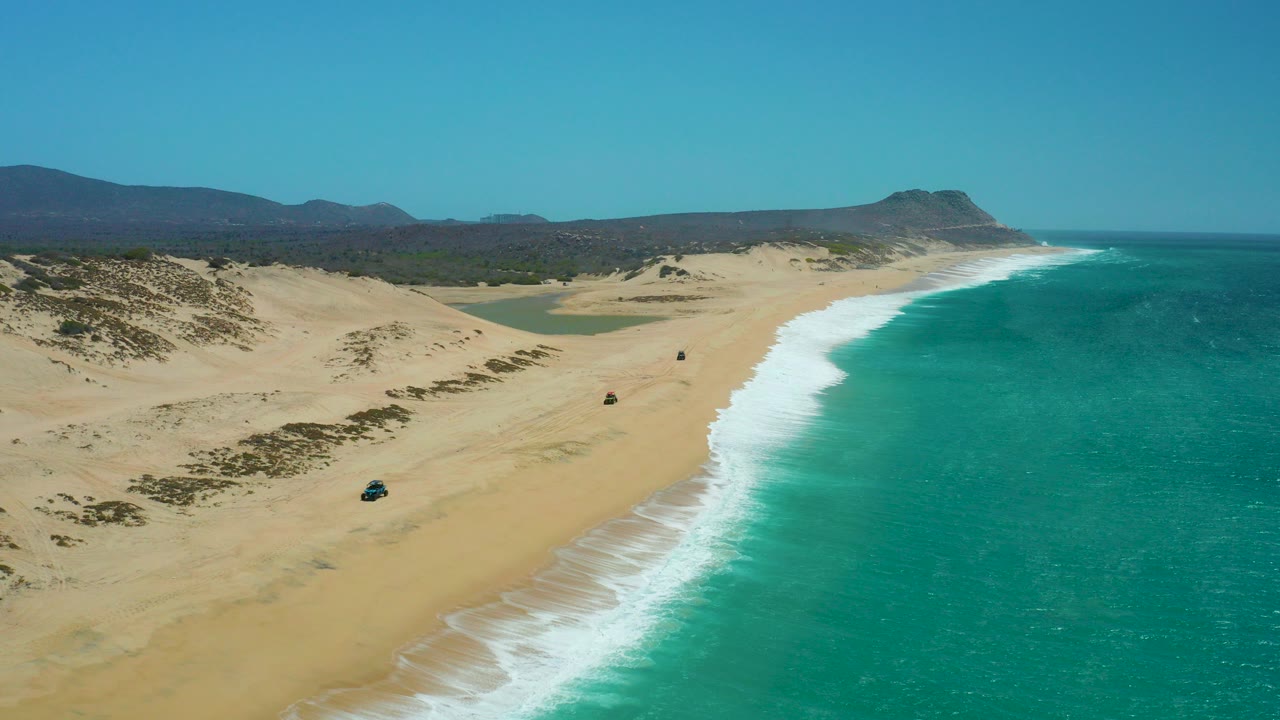 Aerial frame of La Laguna Beach Los Cabos with a small 4x4 low on the sandy beach, dunes to the left, turquoise surf to the right, distant hills, and no visible people.