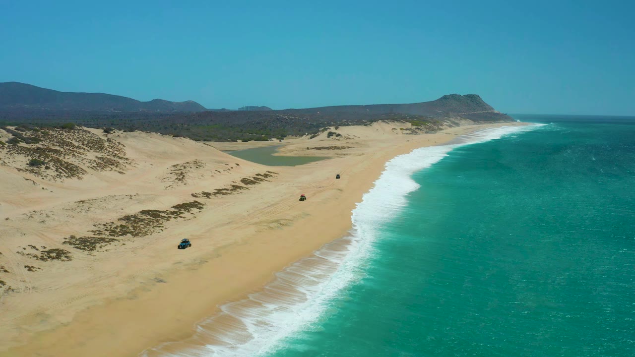 Aerial view over La Laguna Beach Los Cabos as the 4x4 advances along the open sand, with a pale dune field at left, foamy shoreline at right, and no visible people.