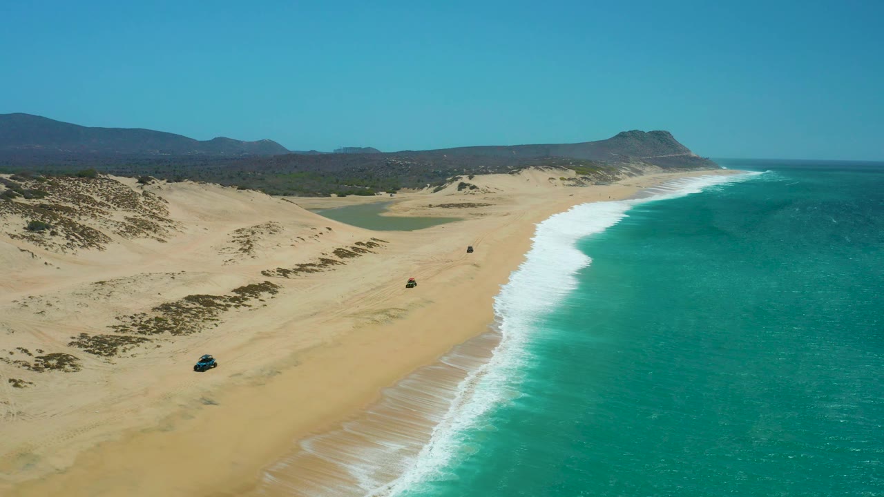 High aerial frame of La Laguna Beach Los Cabos with the 4x4 slightly farther along the sand, white waves pushing up the shoreline, low dunes inland, and no visible people.