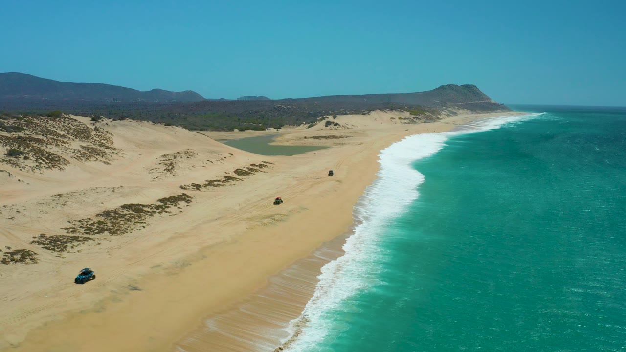 Aerial beach frame with 4x4 near dunes and turquoise surf beside it.