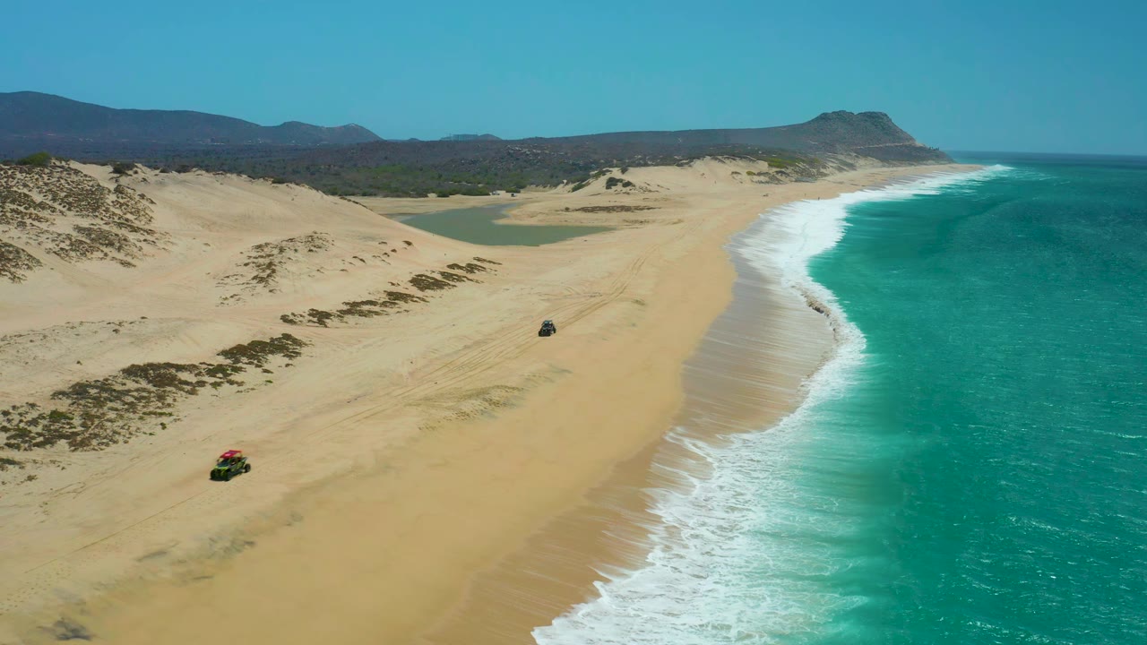 At 0:01, the La Laguna Beach Los Cabos aerial angle keeps both 4x4 vehicles visible on the sand, with one lower left and one nearer center, surf curling along the right edge, and no visible people.