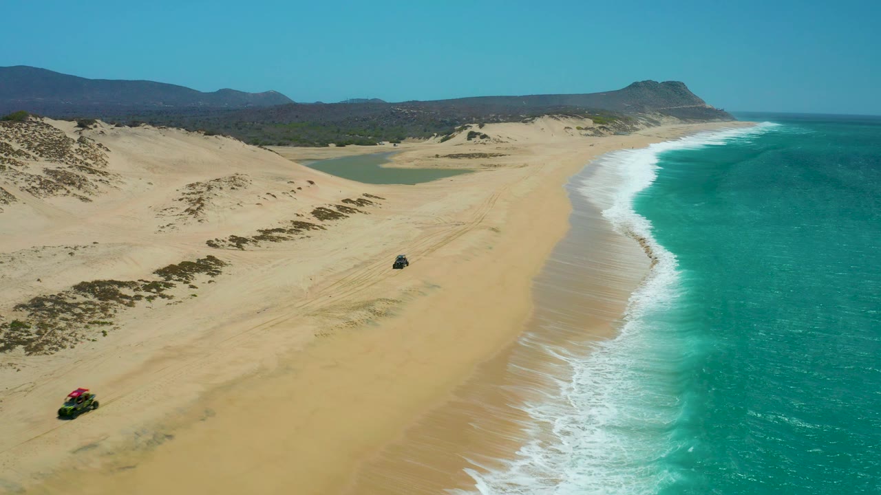 At 0:02, two 4x4 vehicles are farther along the La Laguna Beach Los Cabos sand, with darker dune vegetation at left, a bright white wave line at right, distant hills, and no visible people.