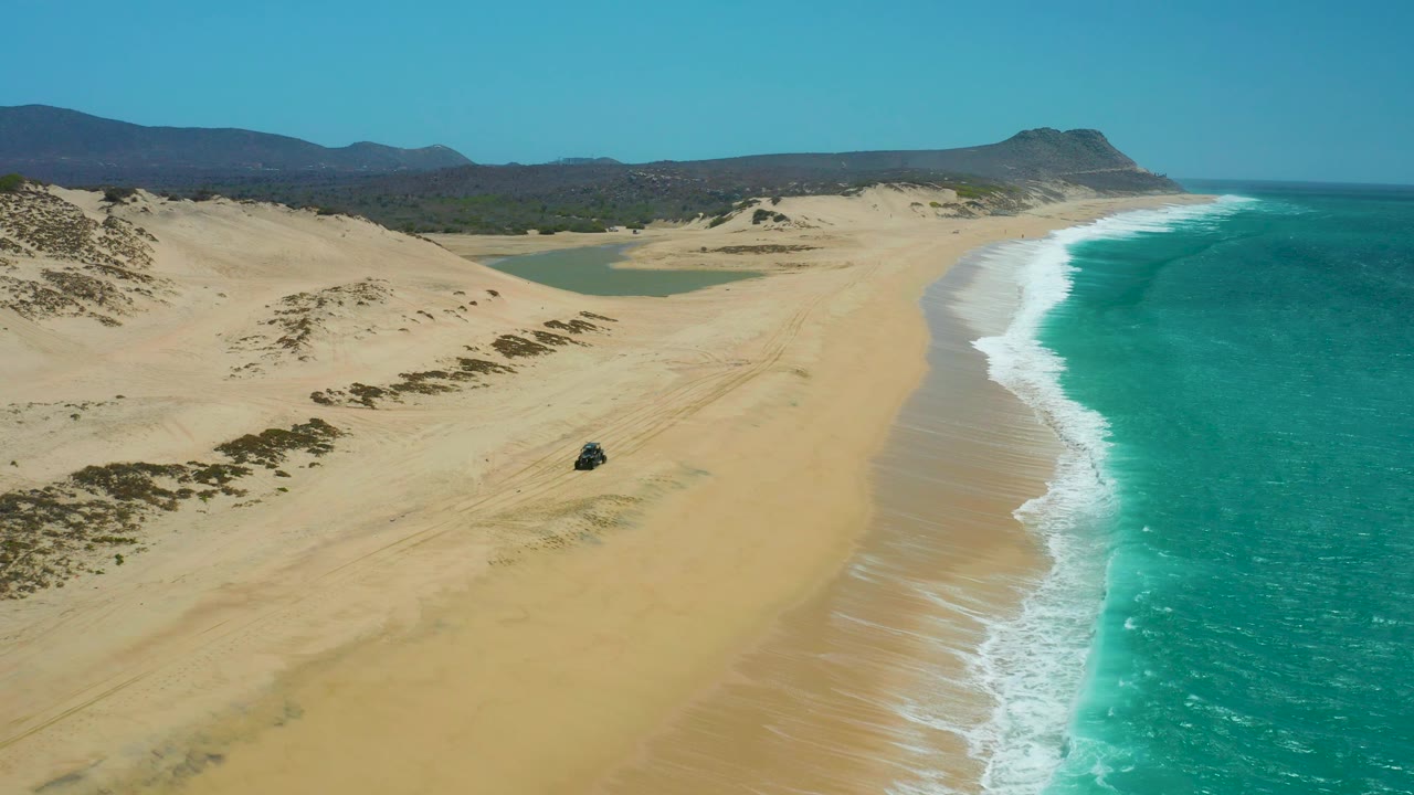 At 0:03, the aerial frame emphasizes the long La Laguna Beach Los Cabos shoreline, with a small 4x4 vehicle on the sand, dunes at left, turquoise water at right, and no visible people.