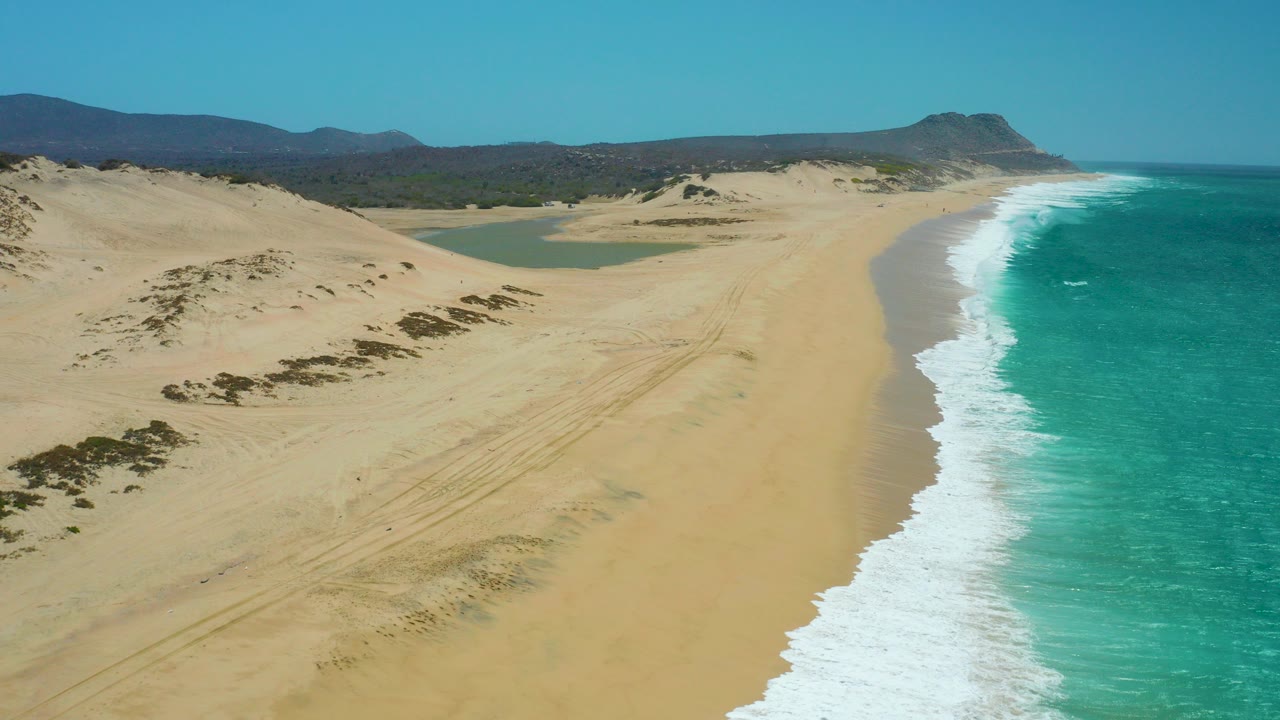 Aerial frame from La Laguna Beach Los Cabos showing turquoise waves on the right, wide sandy beach and dunes on the left, a lagoon inland, distant hills, and no visible people.