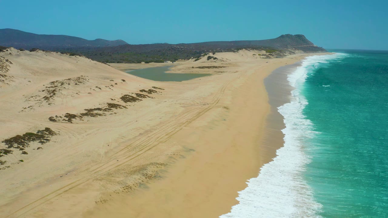 La Laguna Beach Los Cabos appears from above with the surf line slightly farther along the sand, the lagoon set behind the dunes, distant hills on the horizon, and no visible people.