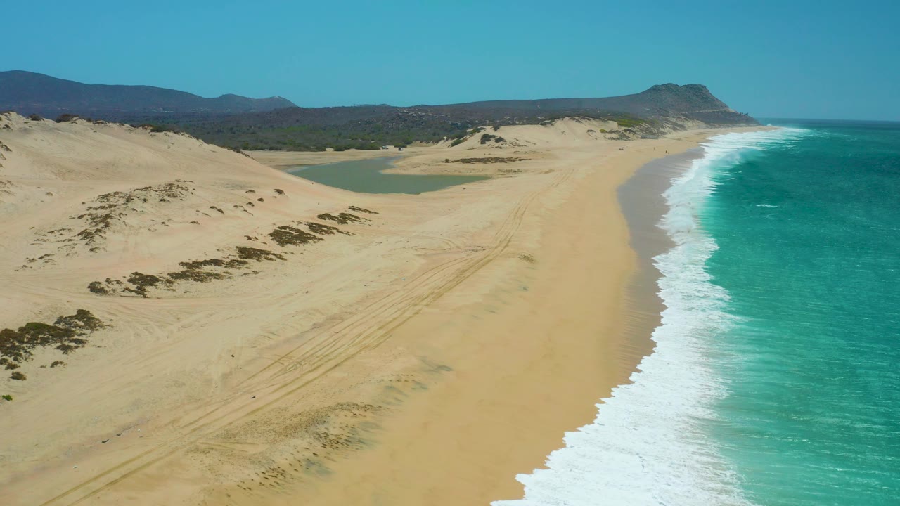 Aerial view of La Laguna Beach Los Cabos with turquoise water filling the right edge, a pale sand shoreline curving into the distance, dunes and lagoon inland, and no visible people.