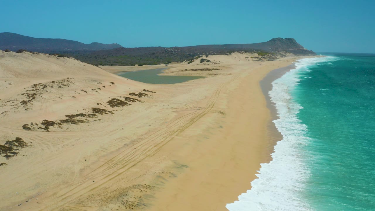 Aerial coastline view with dunes, lagoon, ocean foam, and distant hills.