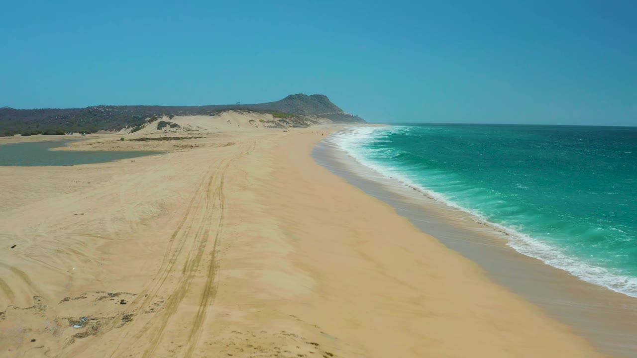 Aerial view at La Laguna Beach Los Cabos showing wide open sand, faint tracks running toward the dunes, turquoise surf along the right edge, and no visible people.
