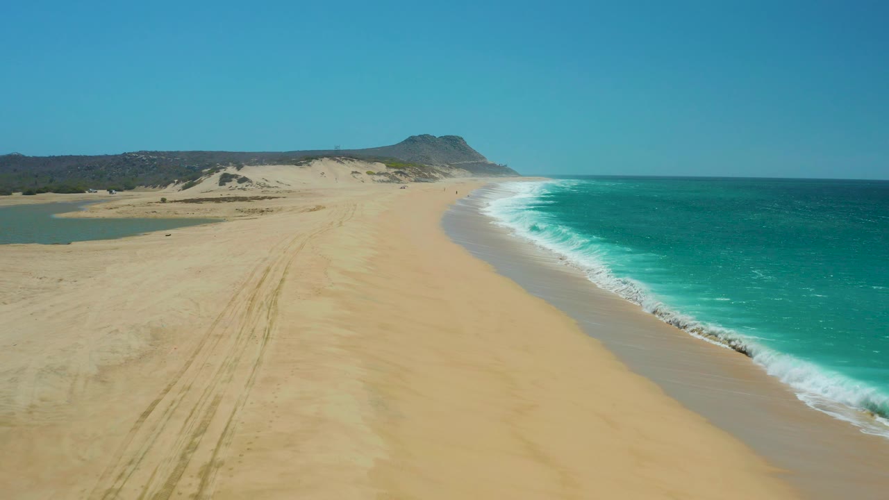 La Laguna Beach Los Cabos aerial frame with a small lagoon visible at left, a long sandy beach through the center, turquoise water at right, and no visible people.