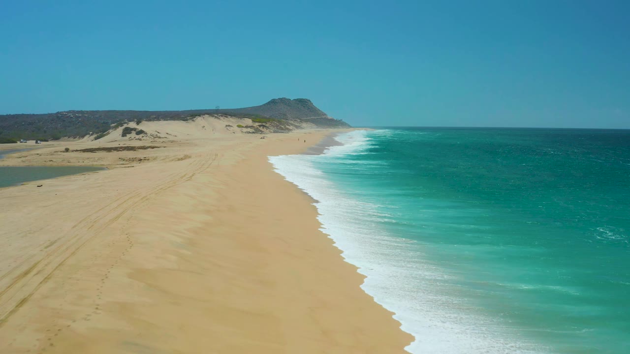 La Laguna Beach Los Cabos shoreline seen from above, with the wave edge slightly farther down the frame, broad sand on the left, teal water on the right, and no visible people.