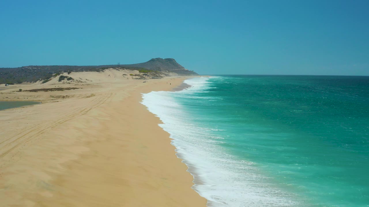 La Laguna Beach Los Cabos appears from a high angle with the surf band lower in frame, uninterrupted sand to the left, open water to the right, dunes behind, and no visible people.