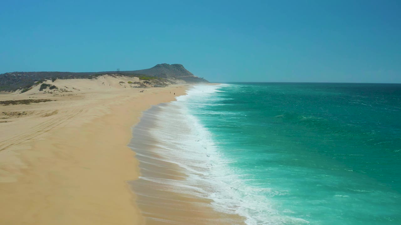 At La Laguna Beach in Los Cabos, a band of white foam runs along the shoreline as blue-green water meets tan sand, with dunes in the background and no visible people.