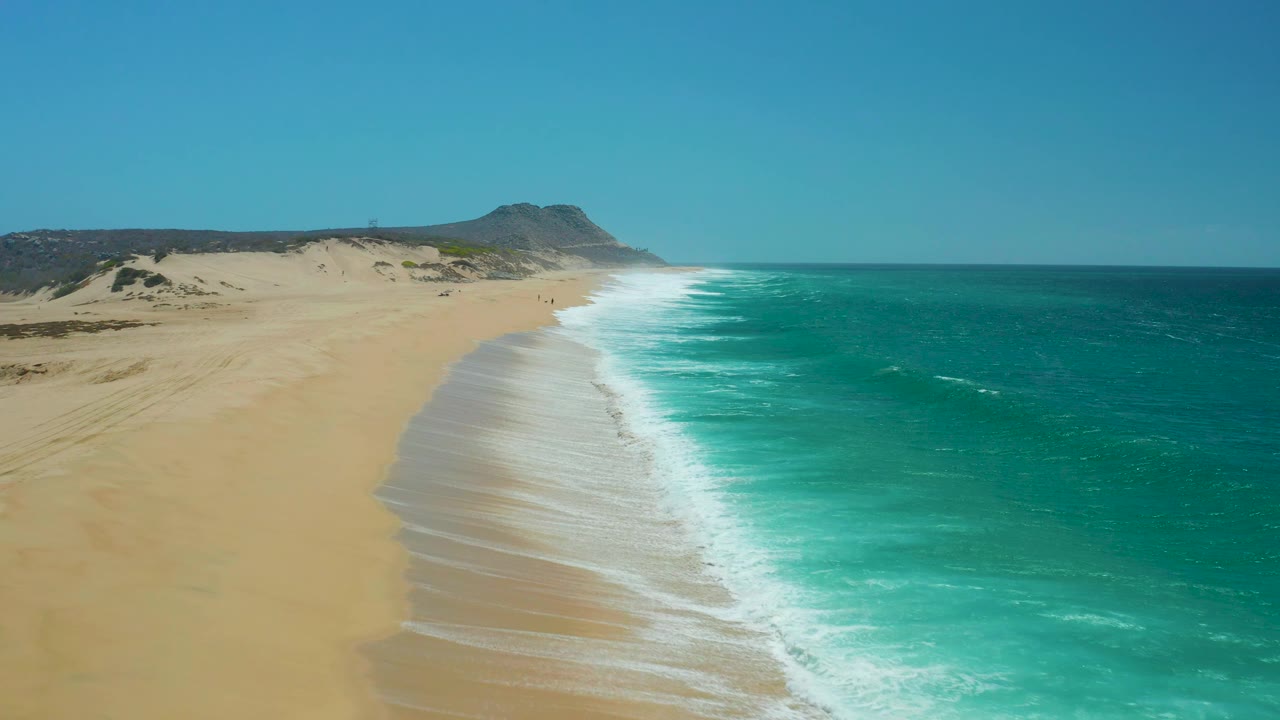 At La Laguna Beach in Los Cabos, rolling surf brightens the right edge of the beach while open sand and low dunes extend toward distant hills, no visible people.