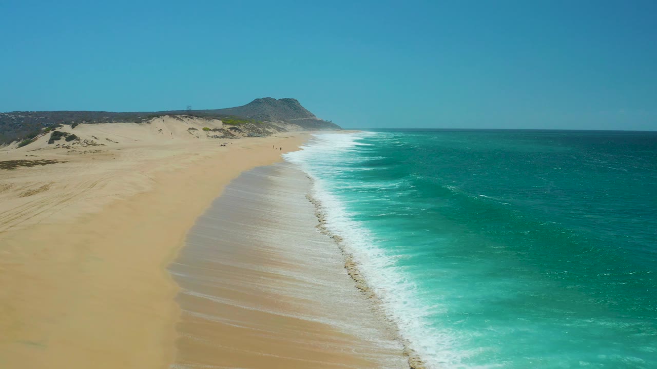 At La Laguna Beach in Los Cabos, the shoreline runs diagonally through the frame with turquoise water on the right, open sand on the left, dunes behind, and no visible people.